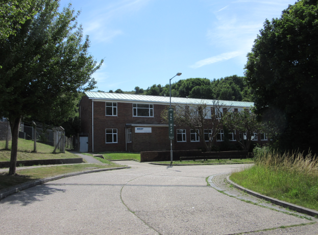 Ein Schulgebäude mit Fenstern, einem Straßenschild, einem Schild, einer Baumgruppe, Gras, einem Zaun und einem bewölkten Himmel, das in der Mitte eines Parks umgeben von saftigem Gras liegt.