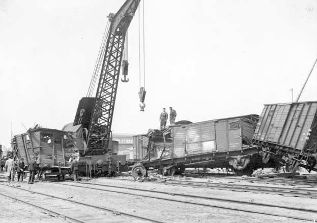 Schwarzes und weißes Bild eines Zugunglücks mit einem Kran, der einen Waggon auf die Schienen hebt, mehrere Menschen in der Nähe, Gebäude und ein klarer Himmel im Hintergrund.