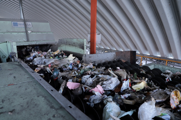 Ein großer Haufen gemischten Mülls auf einem Förderband in einer Recyclinganlage, mit Maschinen und einem Metalldach im Hintergrund.