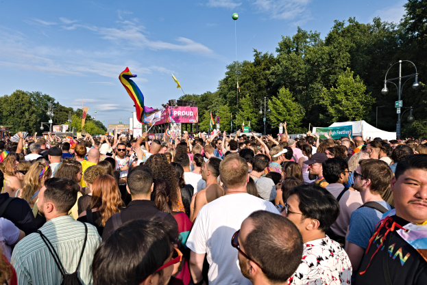 Eine große Menschenmenge steht vor einem Gebäude, einige halten Fahnen und Schilder, mit einem Zelt, Laternenmasten, Bäumen und einem Luftballon im Hintergrund, versammelt zum Feiern der LGBT-Gemeinde.