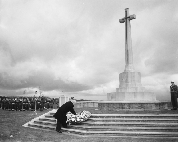 Ein Mann legt einen Kranz auf die Stufen eines Weltkriegsdenkmals, umgeben von einer Gruppe von Menschen, mit Bäumen und einem bewölkten Himmel im Hintergrund, dargestellt in Schwarz-Weiß.