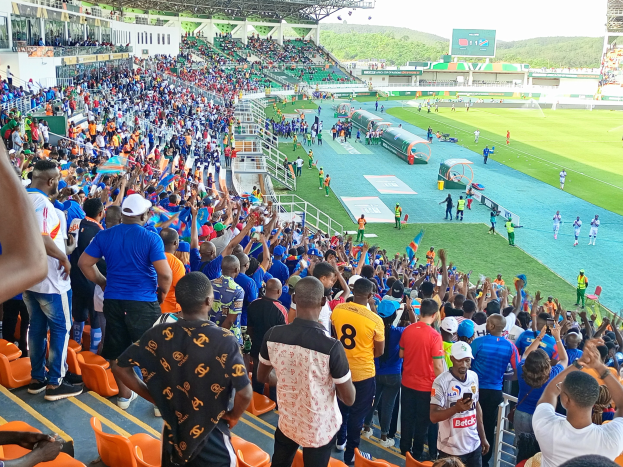Eine große Menschenmenge versammelt sich vor einem eingezäunten Fußballfeld, wobei einige auf Stühlen sitzen und andere stehen, vor einem Hügel und einem klaren blauen Himmel während der Afrikameisterschaft 2019.