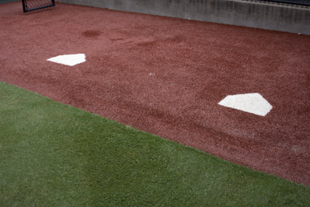 Baseballfeld mit Kunstrasen, umgeben von einem Zaun, mit einem Home Plate in der Mitte und einer Wand im Hintergrund.