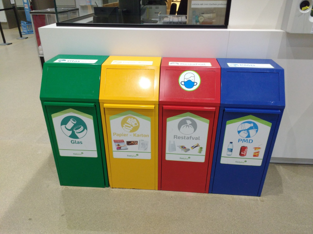 Vier bunte Mülltonnen in einer Reihe auf dem Boden, mit einer Arbeitsplatte, einem Monitor und anderen Gegenständen im Hintergrund und einer Person links daneben.