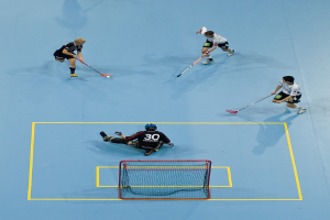 Drei Personen in Sportkleidung spielen Hockey, wobei eine Person rechts mit einem Hockeystock einen Ball schießt und eine Person hinter einem Tor auf dem Boden sitzt.