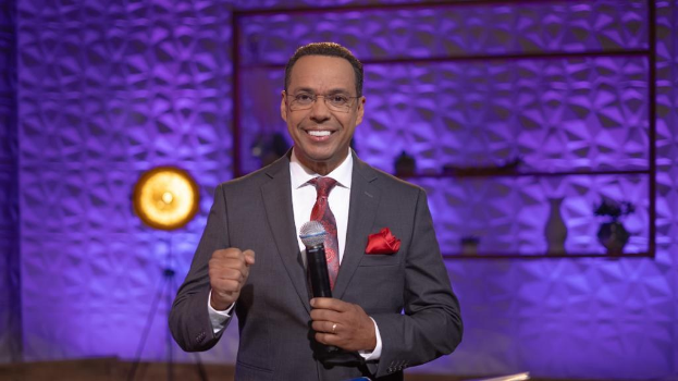 Stephen Colbert, Moderator von *The Daily Show with Trevor Noah*, steht auf einer Bühne und hält ein Mikrofon, trägt einen Anzug, Krawatte und Brille, mit einer Wandmontage und einem Licht hinter ihm.