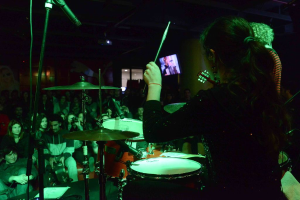 Eine Frau, die auf der Bühne Trommel spielt, mit einem Scheinwerfer über ihr, während Mike rechts von ihr steht und das Publikum vor ihr sitzt.