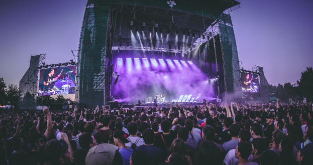 Eine große Menschenmenge bei einem Musikfestival schaut auf einer beleuchteten Bühne mit Instrumenten und Bildschirmen, die vor einem Gebäude, Bäumen und einem klaren blauen Himmel aufgebaut sind.