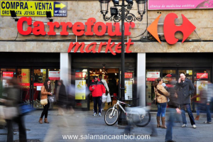 Ein belebter Einkaufsmarkt mit Menschen vor einem Einkaufszentrum, einem schwarzen Pfahl mit Lichtern, einem Fahrrad neben dem Pfahl und einem Richtungsschild an der Wand.