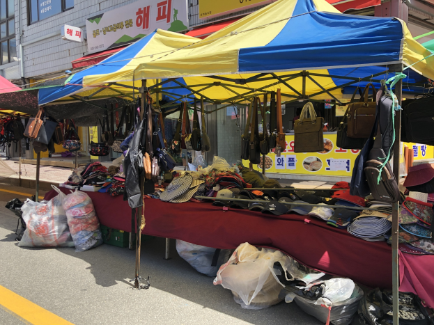 Ein Markt mit Ständen, die Taschen, Hüte und andere Artikel präsentieren, vor einem Hintergrund aus Gebäuden mit Fenstern und Texttafeln.