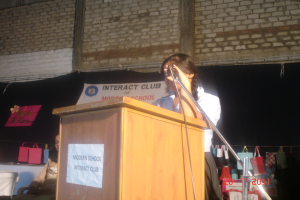 Ein Mädchen in einem weißen Hemd steht in der Nähe eines Rednerpults in einem Konferenzraum, mit einem Mikrofonständer rechts daneben, einem weißen Banner auf einem Hintergrund und mehreren Taschen, die an einem Faden rechts hängen.