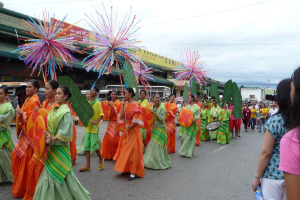 Eine Gruppe von Menschen in farbenfroher Kleidung marschiert während eines Karnevalsumzugs eine Straße entlang, wobei eine Frau auf der rechten Seite eine Flasche hält, und Fahrzeuge, Gebäude, Bäume, Pfähle, Drähte und der Himmel im Hintergrund zu sehen sind.