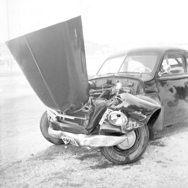 Schwarz-weiß-Foto eines schwer beschädigten Autos am Straßenrand mit eingedellter Motorhaube, zersplitterten Fenstern und offener Fahrertür, die den Motor zeigt.