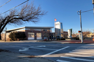 Großes Gebäude mit einer Flagge oben, als neue Feuerwache an einer Straßenkreuzung identifiziert, umgeben von Bäumen, Straßenmöblierung und Infrastruktur, mit Himmel im Hintergrund.