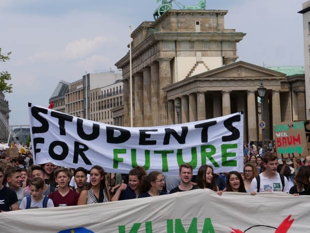 Eine Gruppe von Schülern marschiert in Berlin, eine bunt bemalte Fahne mit der Aufschrift "Students for Future" schwingend, mit Gebäuden, Bäumen und Himmel im Hintergrund.