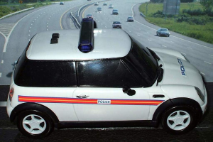 Ein weißer Polizeiwagen steht im Vordergrund, hinter ihm bewegen sich mehrere Autos auf der Straße, umgeben von Gras und Bäumen auf beiden Seiten in einer ländlichen Umgebung.