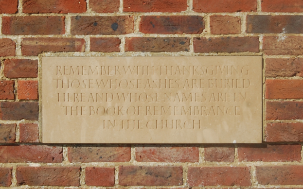 Eine Backsteinmauer mit einer Tafel, auf der steht: "Erinnere dich dankbar an diejenigen, deren Asche hier begraben ist und deren Namen im Buch der Erinnerung in der Kirche stehen."