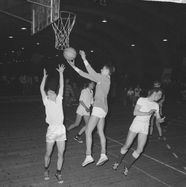 Schwarzes und weißes Foto von jungen Männern, die Basketball in einer Turnhalle mit einem Basketballkorb und Zuschauern im Hintergrund spielen.
