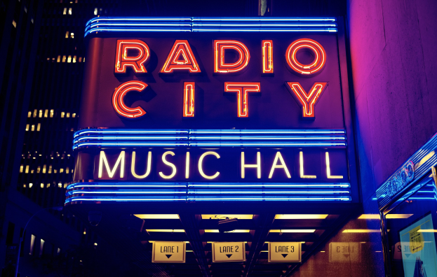 Außenansicht des Radio City Music Hall in New York City bei Nacht, hell erleuchtet mit hohen Gebäuden im Hintergrund und einer 'Radio City'-Leuchtschrift auf der rechten Seite.