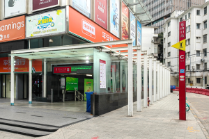Eine Bushaltestelle auf einer Stadtstraße mit Gebäuden auf beiden Seiten, rote und gelbe Schilder mit Text auf der rechten Seite, ein Müllcontainer in der Mitte der Straße, Menschen auf dem Gehweg und eine Rolltreppe auf der linken Seite.