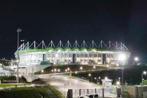 Ein großes Stadion bei Nacht beleuchtet, umgeben von Straßenlaternen, Strommasten, Autos, Grills, Bäumen und einem sternenklaren Himmel.