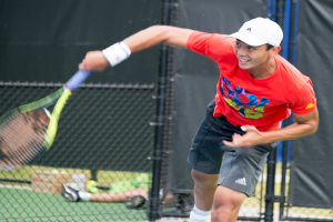 Ein Mann in roter Jacke und weißer Mütze spielt Tennis, hält einen Schläger und hat eine Netz- und durchsichtige Hintergrund; eine weitere Person sitzt in der Nähe auf dem Boden mit einer Box und Bällen.