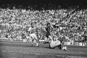 Schwarzes und weißes Foto von Männern, die Fussball auf einem Feld mit Zuschauern im Hintergrund und einem Banner "Bundesliga 1978-79 Ajax 1-1 Hannover 96" spielen.