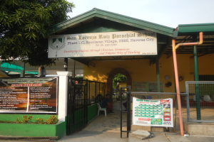 Der Eingang der San Lorenzo Ruiz Pfarrschule ist mit einer Tafel mit Text, einem Metallzaun, Menschen auf Stühlen unter einem Dach, Pfosten, einer Schautafel, Pflanzen, einem Baum und einem bewölkten Himmel versehen.