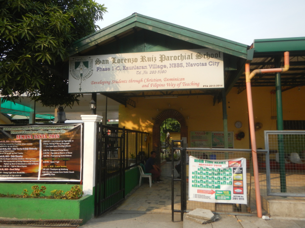 Der Eingang der San Lorenzo Ruiz Pfarrschule ist mit einer Tafel mit Text, einem Metallzaun, Menschen auf Stühlen unter einem Dach, Pfosten, einer Schautafel, Pflanzen, einem Baum und einem bewölkten Himmel versehen.