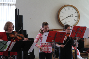 Drei Frauen auf der Bühne mit Geigen spielend, vor sich Notenständer und Noten, während ein Zuschauer im Vordergrund sitzt; eine Uhr hängt an der weißen Wand im Hintergrund, und ein weißer Vorhang ist links sichtbar.