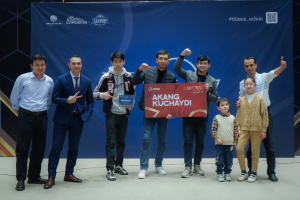 Gruppe von Menschen auf einer Bühne, die eine Tafel mit 'Akang Kuchaydi - Zakovat Quiz' halten, mit einem Banner, Lichtern und anderen Gegenständen im Hintergrund.