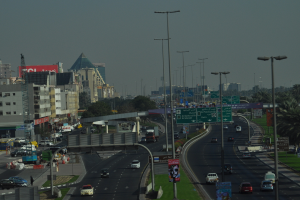 Eine Luftaufnahme einer Stadt mit Gebäuden, Türmen, Fahrzeugen, Werbetafeln, Schildern, Straßen, Bäumen, Straßenlaternen, Menschen, Absperrgittern und Informationsanzeigen unter einem klaren Himmel.