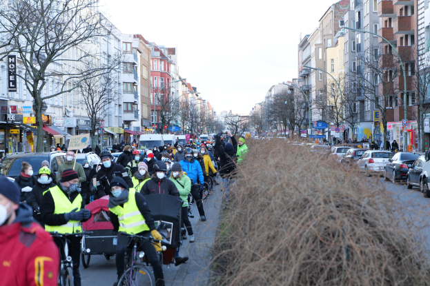 Eine große Gruppe von Menschen in Masken und Sicherheitswesten auf Fahrrädern eine von Bäumen gesäumte Straße entlang, begleitet von Fahrzeugen und trockenem Gras, unter einem klaren blauen Himmel.