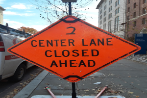 Orange '2 Center Lane Closed Ahead' Schild auf einer Straße mit Fahrzeugen, Polen, Brettern, Bäumen, Gebäuden und einem bewölkten Himmel im Hintergrund.