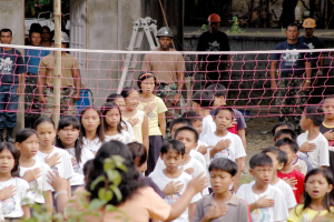 Eine Gruppe von Kindern steht vor einem Netz mit einer Frau im Vordergrund, während Menschen mit Helmen, eine Leiter, eine Wand und Bäume im Hintergrund zu sehen sind.