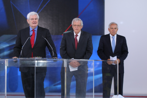 Ein älterer Mann in einem schwarzen Mantel und roter Krawatte steht an einem durchsichtigen Rednerpult auf einer Bühne, mit einem blauen und weißen Banner im Hintergrund.
