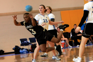 Mehrere Mädchen spielen mit einem blauen Ball auf einem Indoor-Court, mit Stühlen, Gepäcktaschen und anderen Menschen im Hintergrund.
