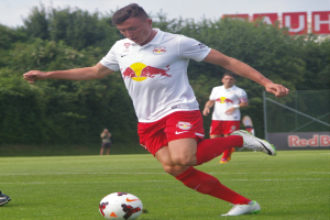 Ein Mann in einem Fussballtrikot schie├čt einen Ball auf einem grasbewachsenen Feld mit B├Ąumen und einem klaren blauen Himmel im Hintergrund, wobei ein "RB Leipzig"-Schild zu sehen ist.