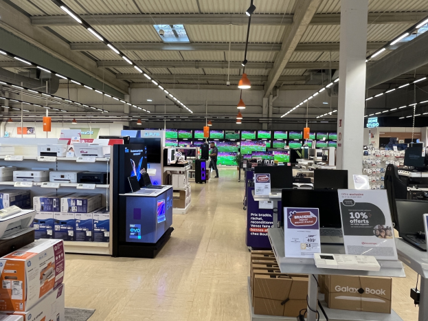 Ein großer Raum in einem Walmart-Laden voller Elektronik, die in Regalen angeordnet ist, einschließlich Computer und Drucker, mit Menschen auf dem Boden, einer Säule, Schildern, Deckenleuchten und Deckenventilatoren.