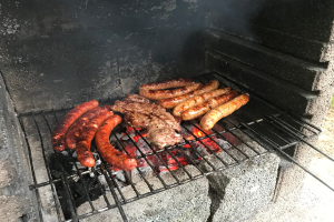 Grill mit Würstchen und Fleisch darauf, umgeben von brennender Kohle und Flammen, im Freien auf Gras stehend.