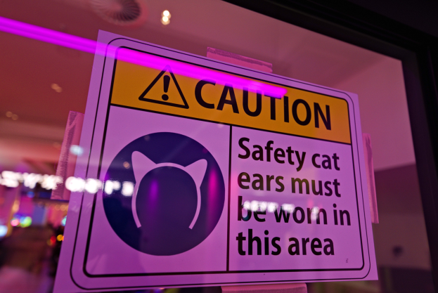 Hellgelbes Warnschild in einem Schaufenster mit der Aufschrift "Vorsicht: Sicherheitskatzenohren müssen in diesem Bereich getragen werden."