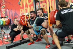 Eine Gruppe von Männern führt Kniebeugen mit Hanteln in einem Fitnessstudio durch, das mit Geräten und Bannern ausgestattet ist, auf denen "Powerlifting-Weltmeisterschaften 2019" steht.
