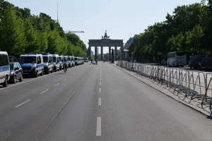 Eine lange Reihe von Polizeiwagen, die auf der Seite einer Straße vor dem Brandenburger Tor in Berlin, Deutschland, geparkt sind, mit Menschen, die Fahrräder fahren und auf der Straße stehen, sowie Absperrungen und Bäume an den Seiten und einem Bogen mit Statuen im Hintergrund.