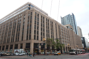 Eine Straße mit Menschen, die Fahrräder, Busse und Autos fahren; im Hintergrund sind Gebäude, ein Baum, Laternen, Pfosten, Drähte und der Himmel zu sehen.