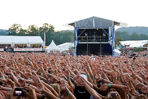 Eine große Menschenmenge auf einem Musikfestival mit erhobenen Händen, einige halten Handys, vor einer Bühne mit Performern, weißen Zelten, Bäumen und einem klaren blauen Himmel.