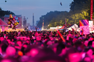 Eine große Menschenmenge bei einem nächtlichen Musikfestival, beleuchtet von bunten Lichtern, umgeben von Zelten, Fahnen, Pfählen und anderen Gegenständen und im Hintergrund ein Turm, Bäume und einen klaren Nachthimmel.