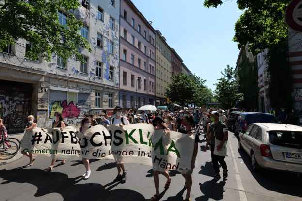 Eine Gruppe von Menschen marschiert mit einem Transparent die Straße entlang, Autos parken rechts, ein Fahrradfahrer links, Gebäude und Bäume im Hintergrund und eine Berliner Demonstration.
