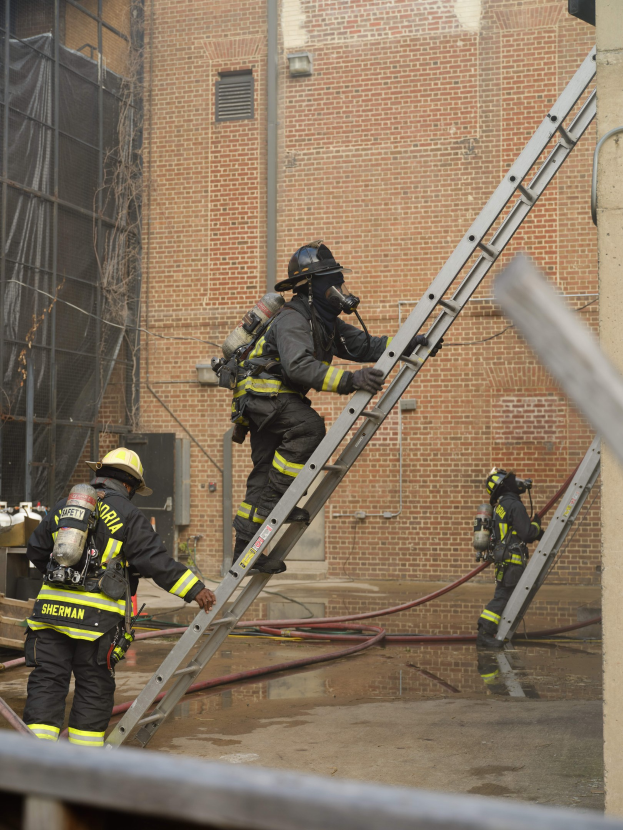 Feuerwehrleute mit Helmen und Lufttanks klettern an einer Leiter vor einem Backsteingebäude mit Rohren auf dem Boden und einer Metallstange am unteren Ende, mit einem weiteren Gebäude und einem Netz im Hintergrund.