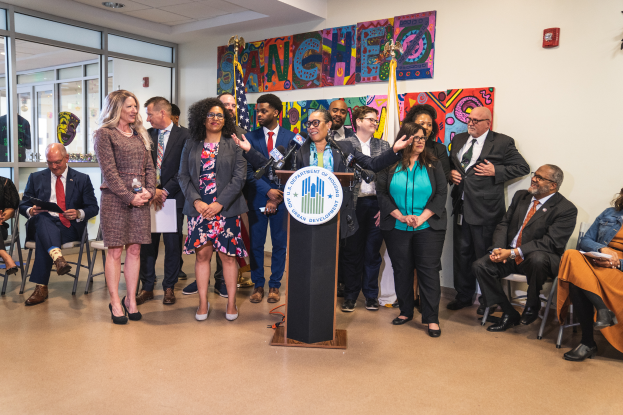 Eine Gruppe von Menschen, die vor einem Podium mit Mikrofon und einer Tafel mit der Aufschrift "Sancho City Councilman Joins City Council" stehen und sitzen, mit Flaggen, einer Tafel, einem Feuerlöscher und Deckenlampen an der Rückwand.