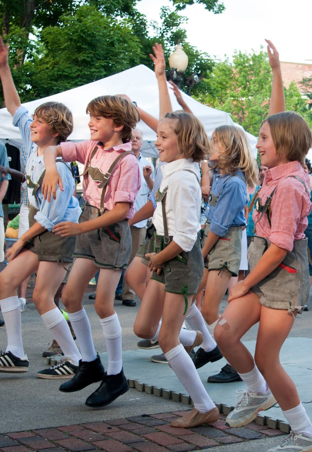Kinder in traditioneller bayrischer Tracht tanzen auf der Straße, umgeben von einer Menge, Zelten und Gebäuden unter einem klaren blauen Himmel.